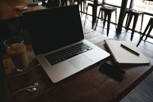 A table at a coffee shop with an iced coffee, an open laptop with earbuds plugged in, a notebook with a pen, and a cell phone